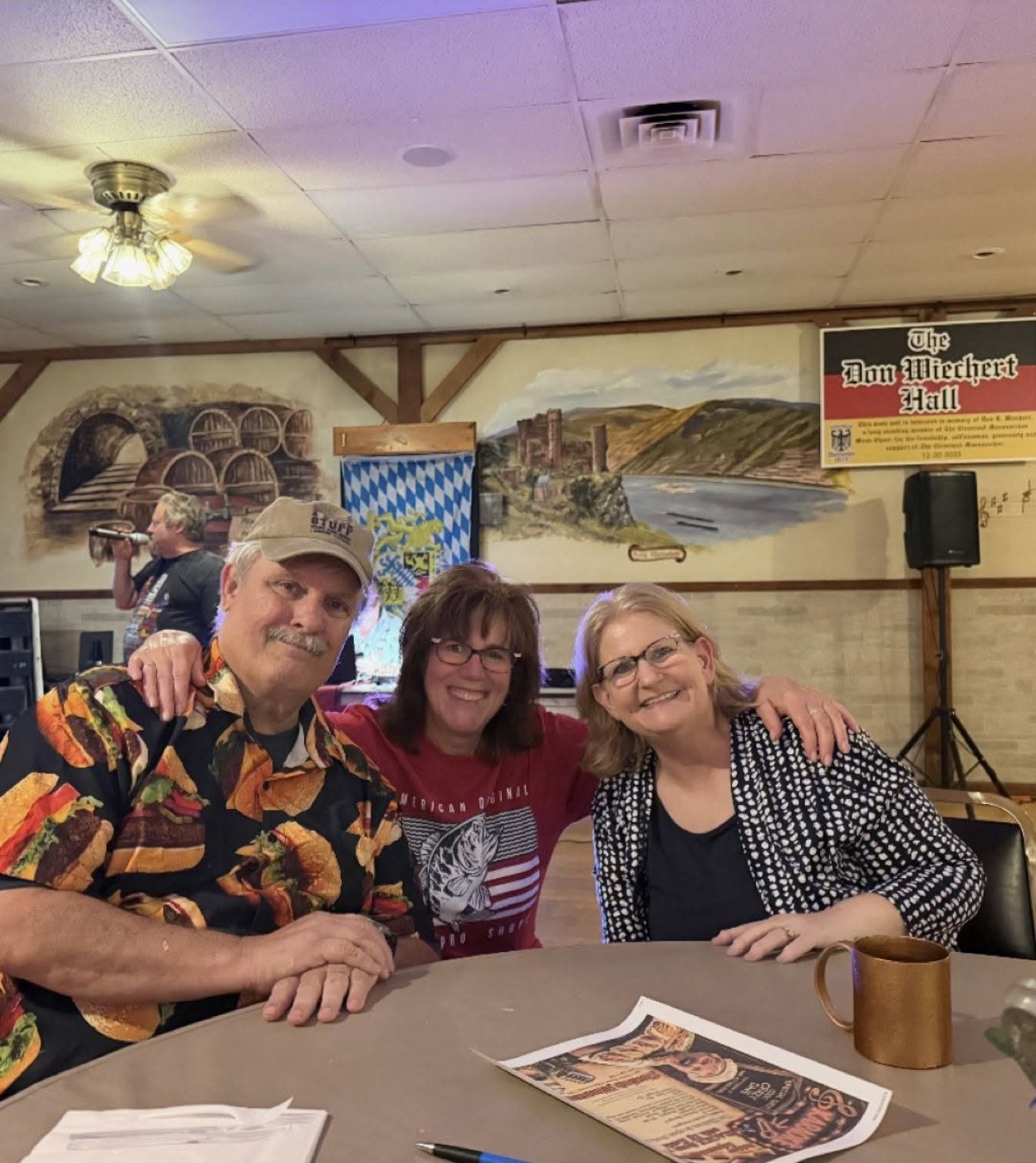 Three people seated at a table in the Don Wiechert Hall at Cleveland Maennerchor, one wearing a burger-print shirt, with the Bavarian murals behind them and the karaoke flyer on the table
