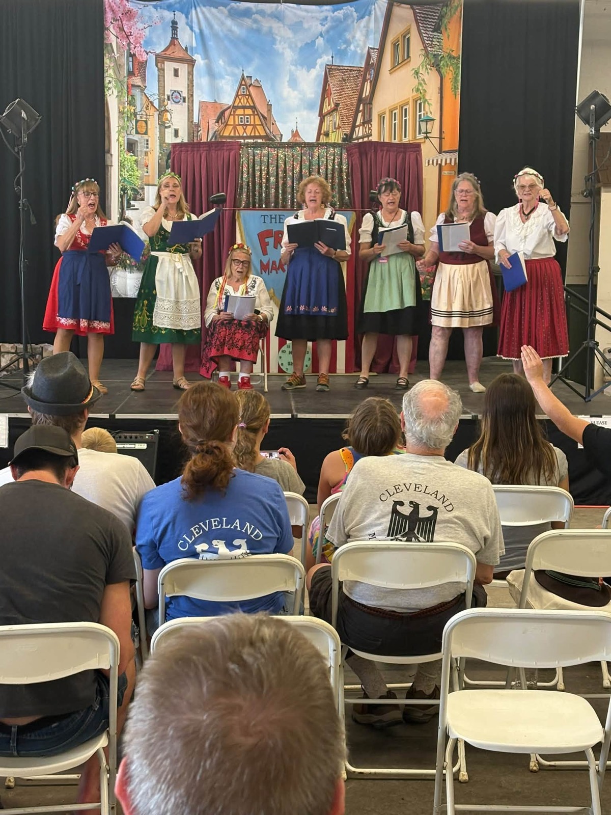 The Cleveland Maennerchor Women's Choir performing on stage at Cleveland Oktoberfest in Berea, singing from folders in front of a painted Bavarian streetscape
